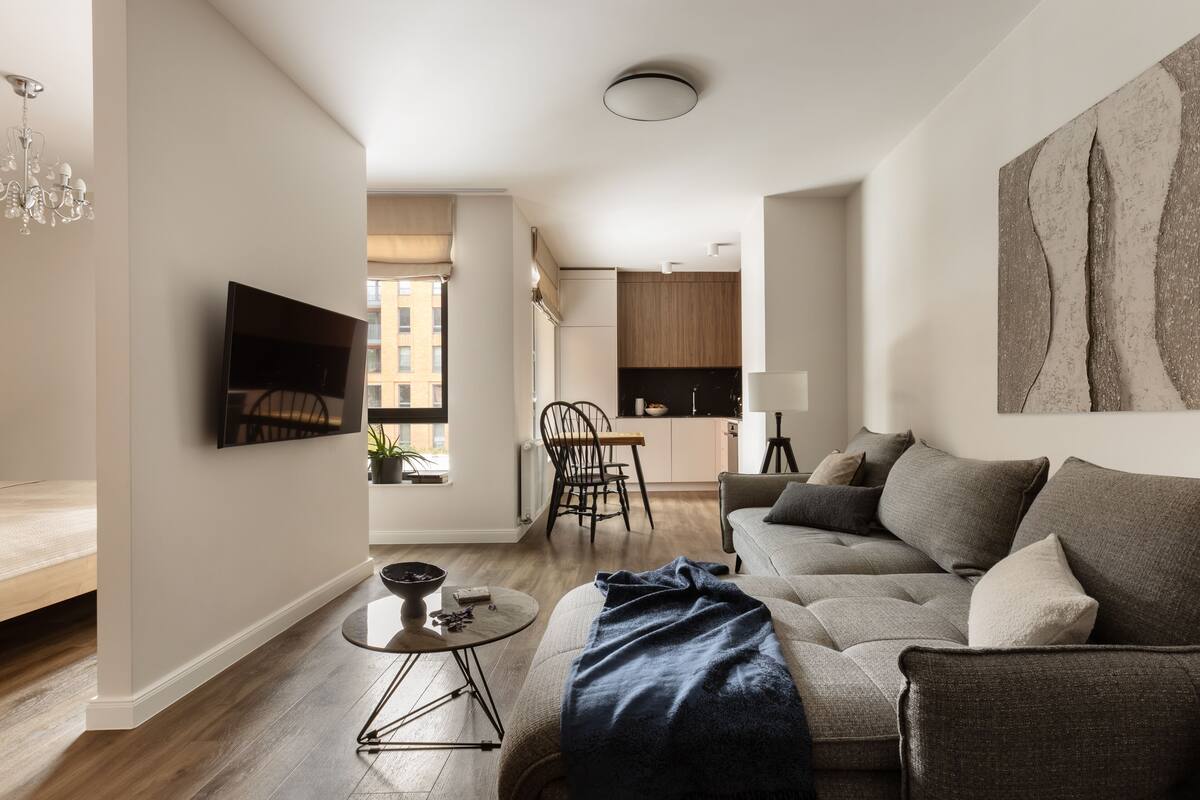 A modern living room with a sofa, a coffee table, and a TV mounted on the wall. There is a kitchen area in the background with cabinets and a dining table.