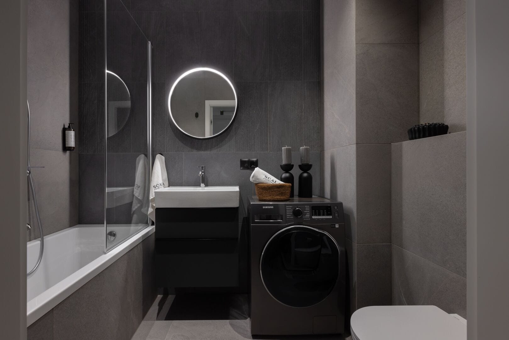 Un baño moderno con bañera, lavabo, lavadora y espejo con luz circular.