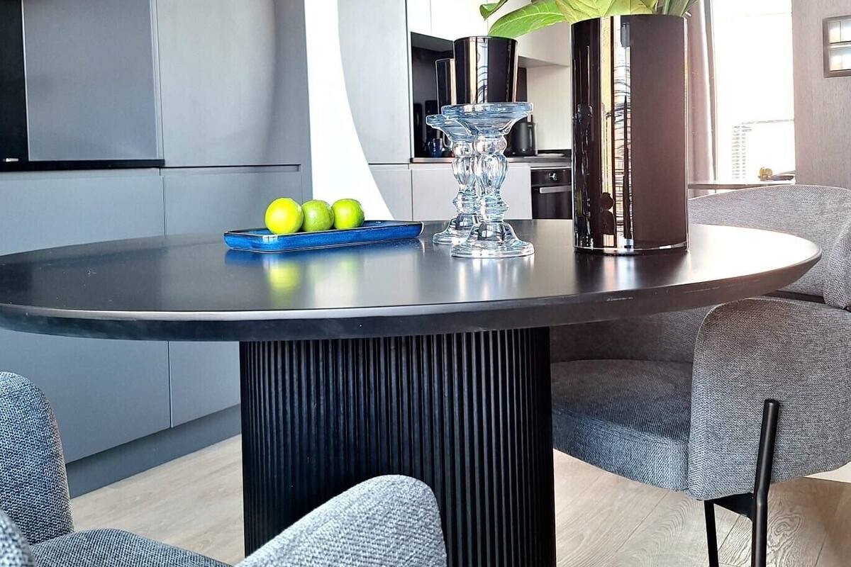 A modern kitchen with a curved island, a dining area with grey upholstered chairs, and a tall black vase with green leaves.