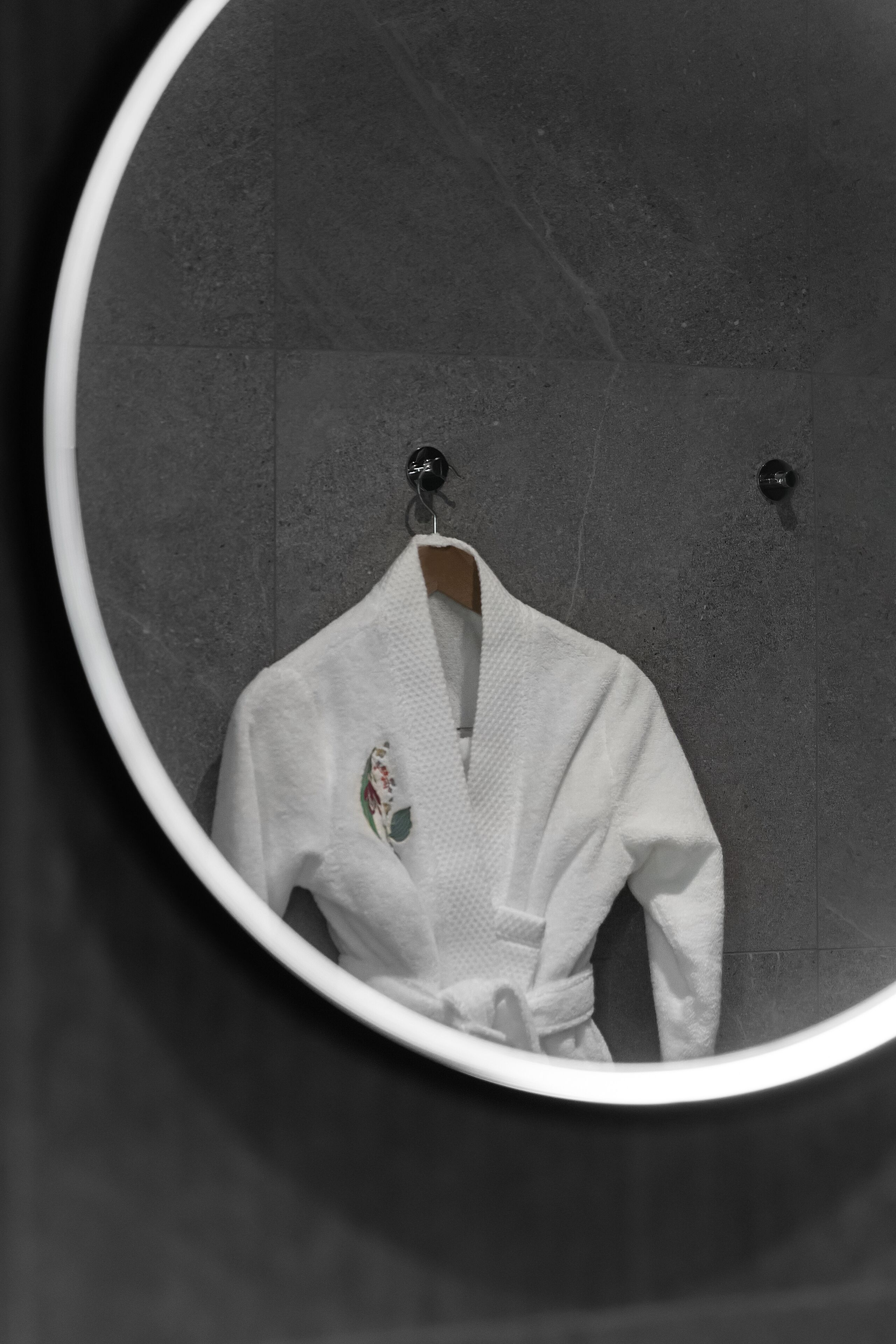 A white bathrobe hanging on a hook in a bathroom with a round mirror.