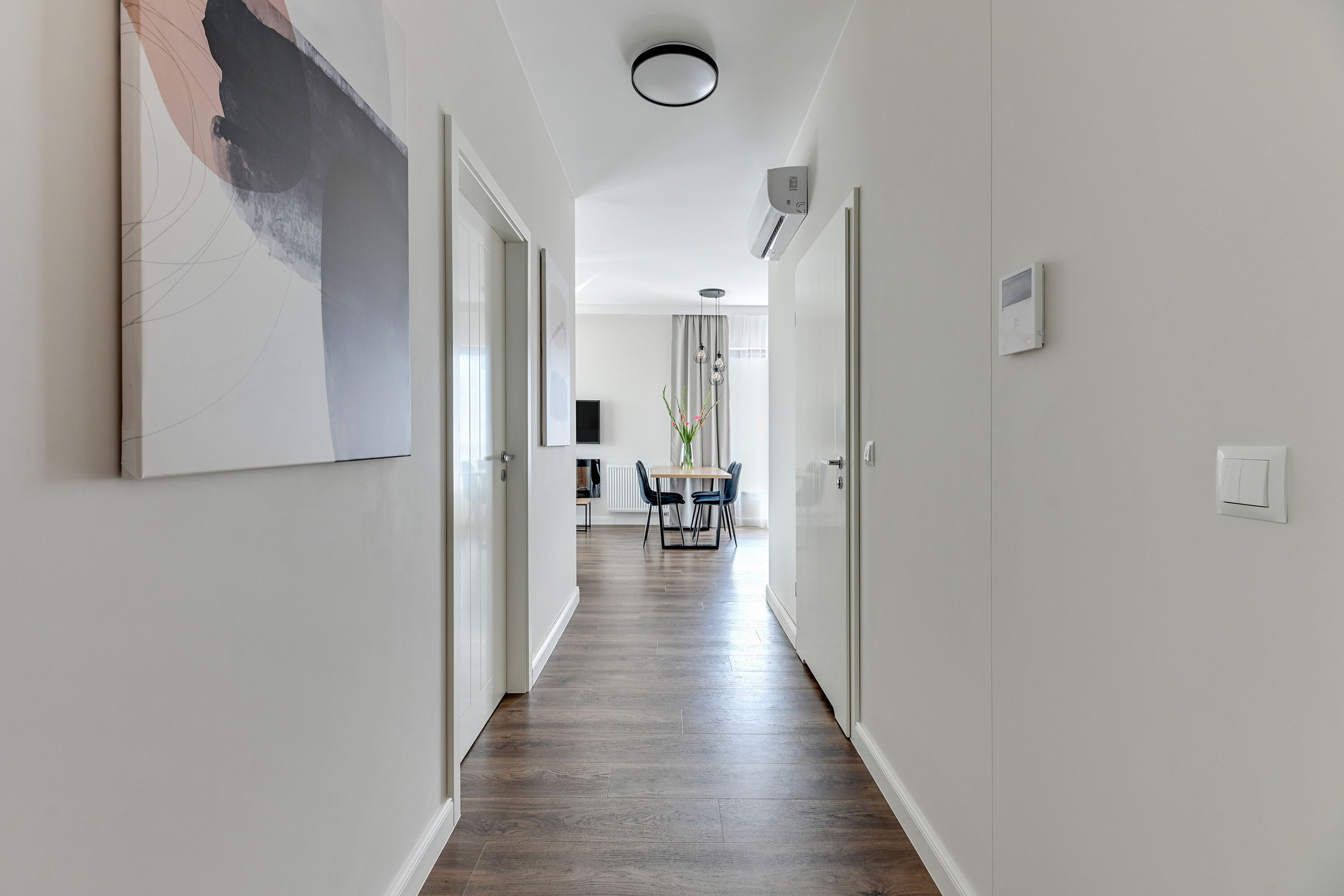 A hallway with a wooden floor, white walls, and a dining area in the distance.
