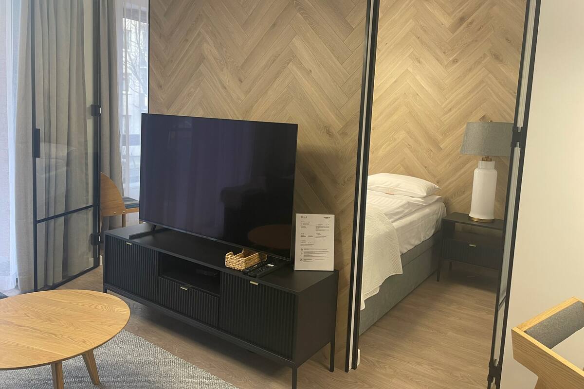 A modern hotel room with a large flat-screen TV on a black entertainment unit, a bed with white bedding, a bedside table with a lamp, and a wooden floor with a herringbone pattern.