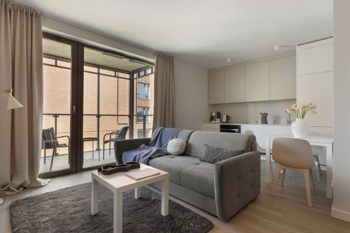 A modern living room with a grey sofa, a small white table, and a black rug. There is a sliding glass door leading to a balcony with chairs.