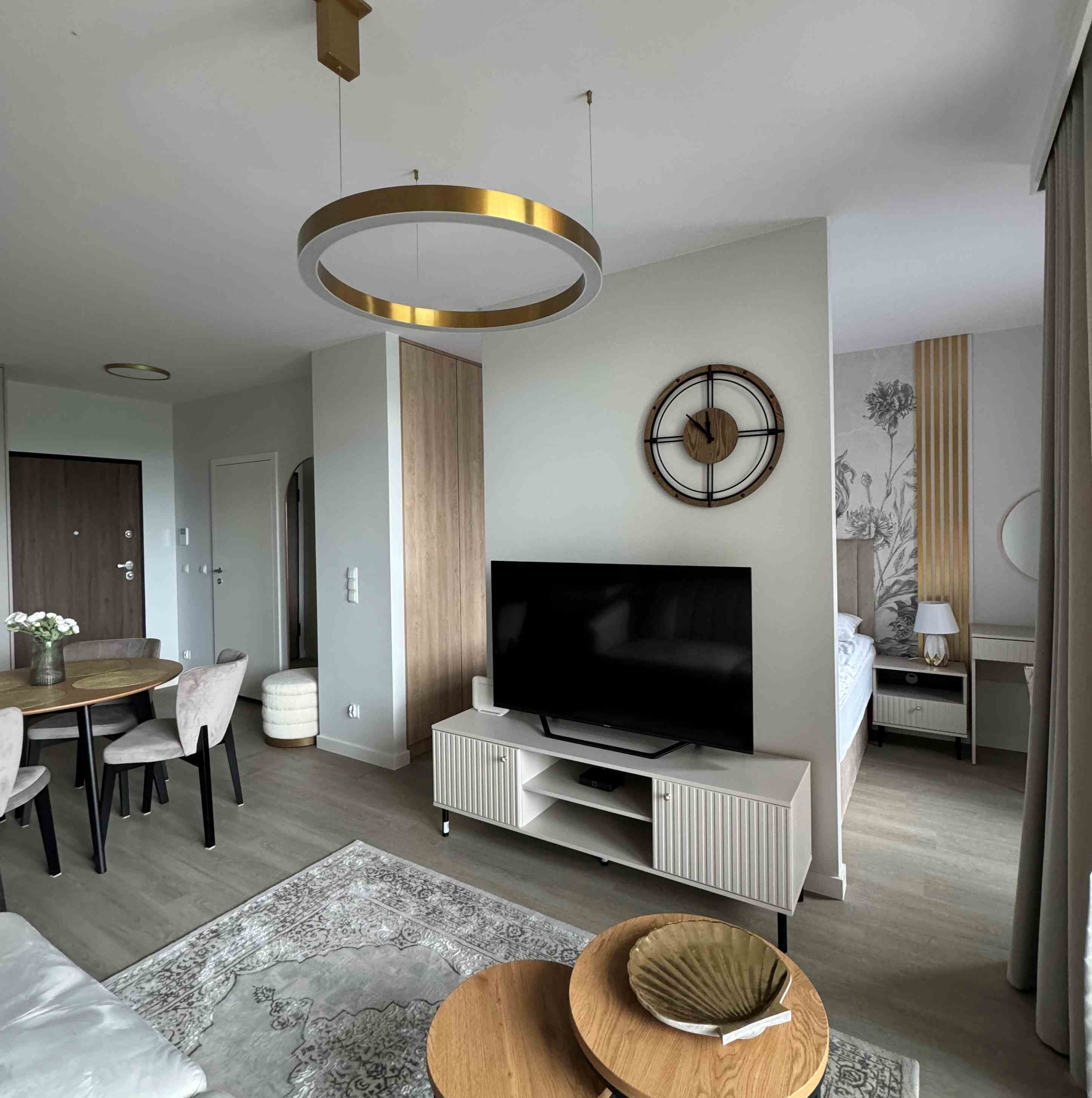 Una sala de estar moderna con un televisor de pantalla plana, un mueble de entretenimiento de madera, una zona de comedor con una mesa y sillas, y un reloj decorativo en la pared.