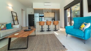Una sala de estar moderna con un sofá, una mesa de centro y un sillón azul. Se observa una zona de cocina con un taburete de bar y una lámpara colgante.