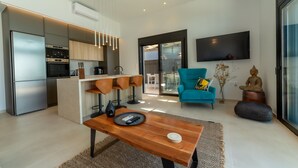Cocina moderna con horno integrado y refrigerador de acero inoxidable. Hay una zona de comedor con mesa y sillas de madera. Un televisor de pantalla plana grande está montado en la pared.