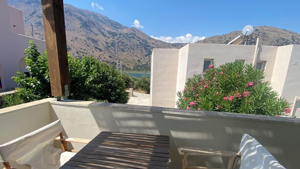 Een balkon met een houten tafel en twee stoelen, met uitzicht op een bergachtig landschap en een wateroppervlak.