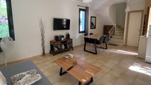 Een ruime woonkamer met een bank, een salontafel, een televisie en een eettafel met stoelen. Er is een trap die naar een hoger gelegen verdieping leidt.