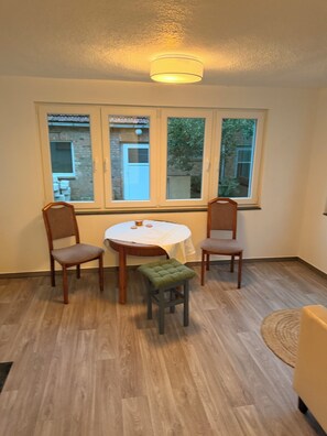 Una habitación con una mesa redonda, dos sillas de madera, un taburete pequeño y una ventana que ofrece vistas a un edificio de ladrillo y vegetación.