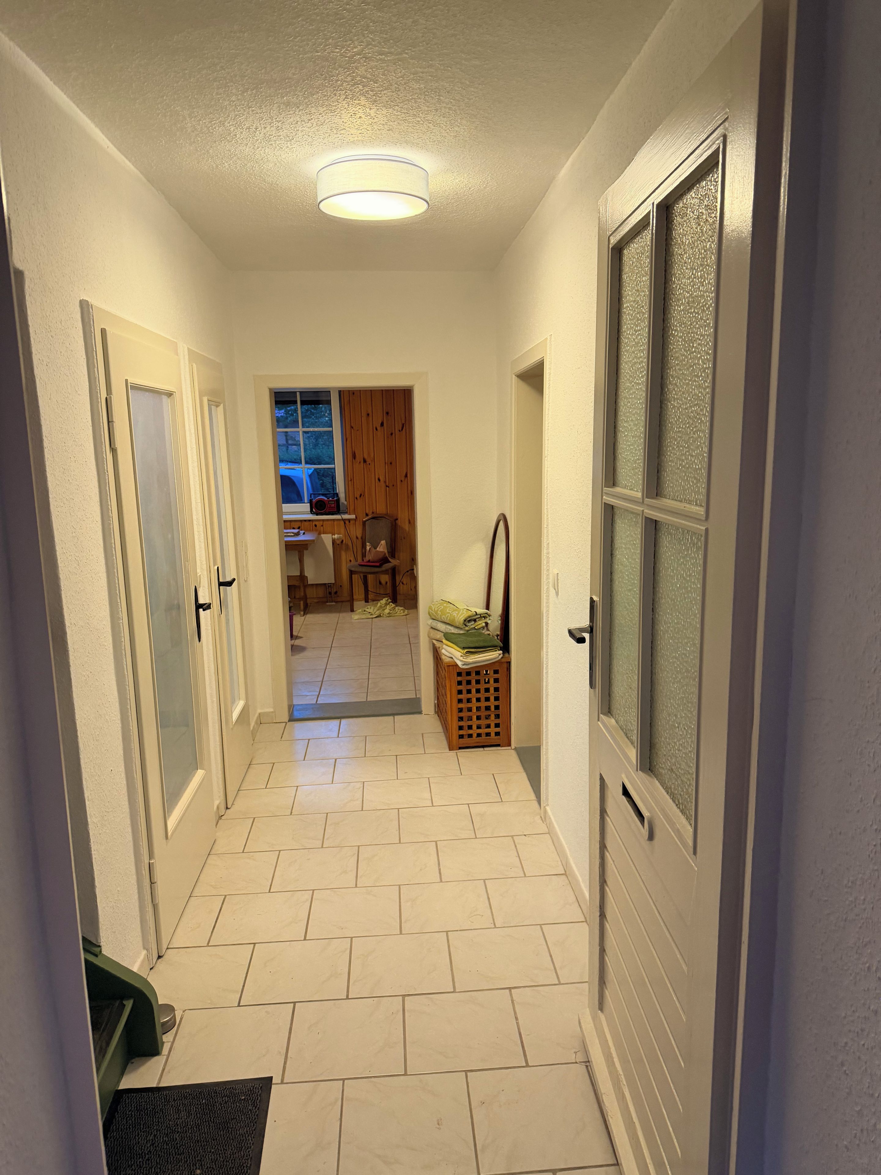 A hallway with white tiled flooring, a ceiling light, and doors leading to other rooms.