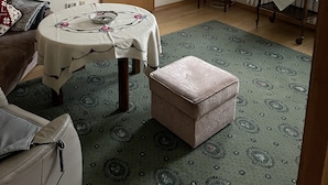 Una sala de estar con una televisión, una mesa con mantel floral, un sofá, una puerta de madera y una alfombra estampada.