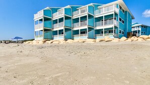 A multi-story beachfront building with balconies, a sandy beach in the foreground, and a clear blue sky.