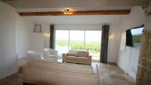 En rummelig stue med en stor beige sofa, en hvid lænestol og et lille hvidt bord. Der er et stort vindue med gardiner, en stenvæg og et ophængt fjernsyn.