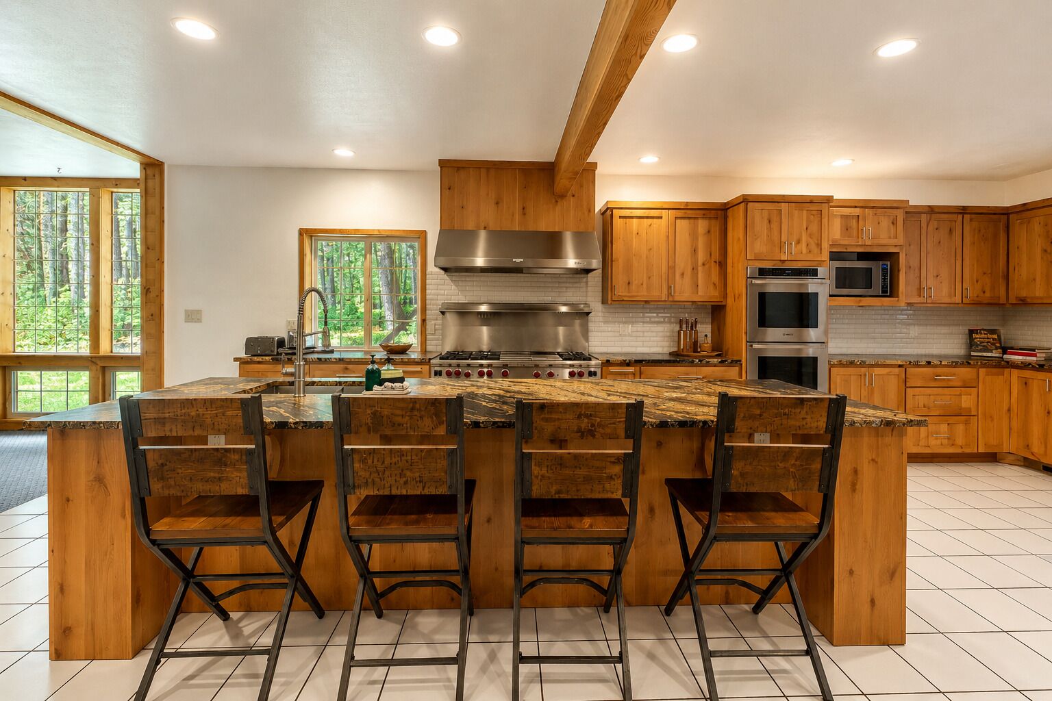 A modern kitchen with wooden cabinets, a central island with bar stools, stainless steel appliances, and large windows.