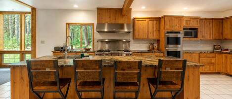 A modern kitchen with wooden cabinets, a central island with bar stools, stainless steel appliances, and large windows.