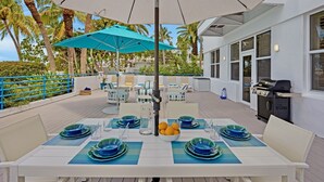 An outdoor dining area with a table set for four, surrounded by chairs, a blue umbrella, and a grill. - **MODERN MAJESTY @ THE ANNEX** Condo & Pool + Last Key White Glove Service... (Key West)