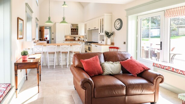 A spacious living room with a brown leather sofa, a wooden coffee table, and a dining area with white chairs and a kitchen in the background. - No. 29 Ness (Isle of Lewis)