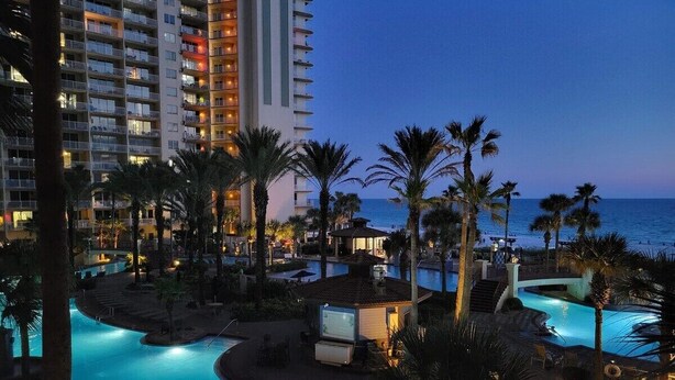 Zona de piscina en un resort con vista al océano y un alto edificio de hotel al fondo.