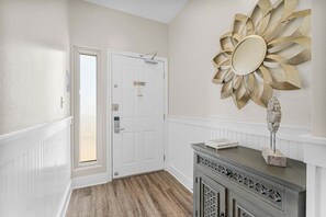 A hallway with a white door, a decorative sun-shaped mirror, and a cabinet with a stone sculpture. - Real Shore 1625–Ocean Views–Sleeps 6 (Panama City Beach)