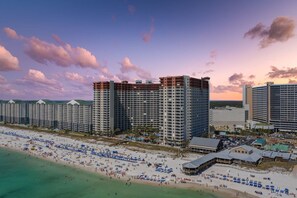 A beachfront hotel with multiple high-rise buildings, a sandy beach with umbrellas, and a clear sky.