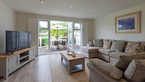 Un salon moderne doté d’un téléviseur à écran plat, d’une table basse en bois, d’un canapé beige et d’une grande porte-fenêtre coulissante donnant sur une terrasse extérieure.