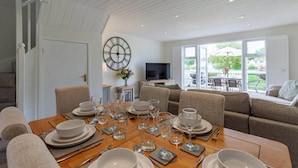 Un coin repas avec une table dressée pour six personnes, une grande horloge murale et un téléviseur en arrière-plan.