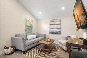 Ein Wohnzimmer mit einem grauen Sofa, einem Holztisch, einem Teppich mit ZEBRA-Muster, einem Fernseher und einem Fenster mit Jalousien.