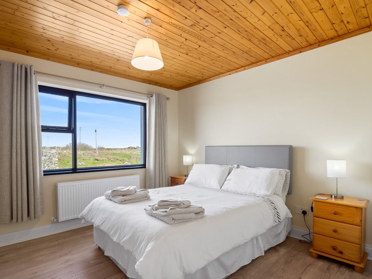 A bedroom with a large bed, a window with curtains, a bedside table with a lamp, and a wooden ceiling.