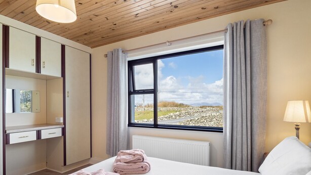Makuuhuoneessa on sänky, työpöytä, ikkuna, jossa on verhot, ja ikkunasta avautuu näkymä kivikkoiseen maisemaan.
