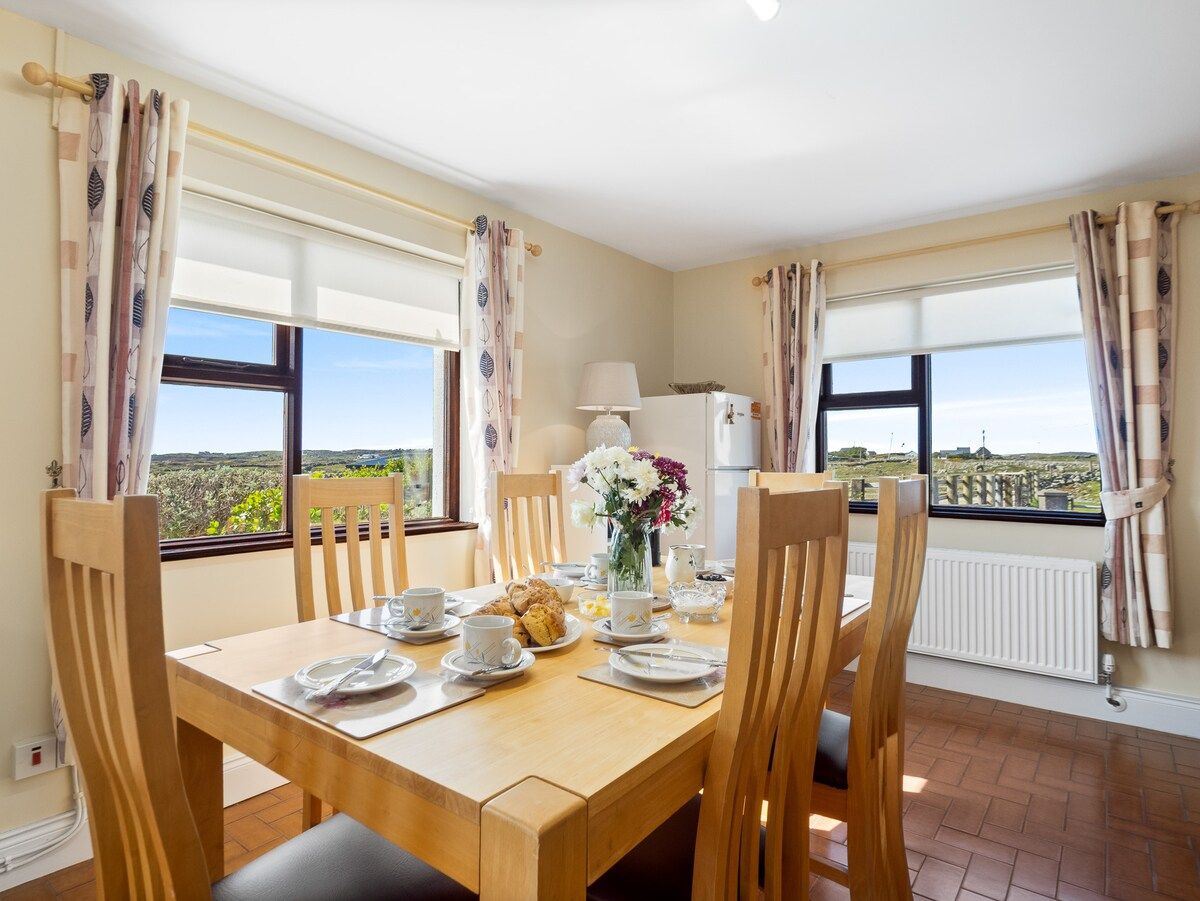 A dining area with a wooden table set for four, chairs, and a view of the countryside through large windows.