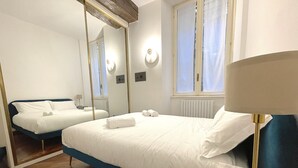 A hotel room with two beds, a large mirror, a bedside table with a lamp, and a window with curtains. - Tower Gombito's Apartment (Bergamo)