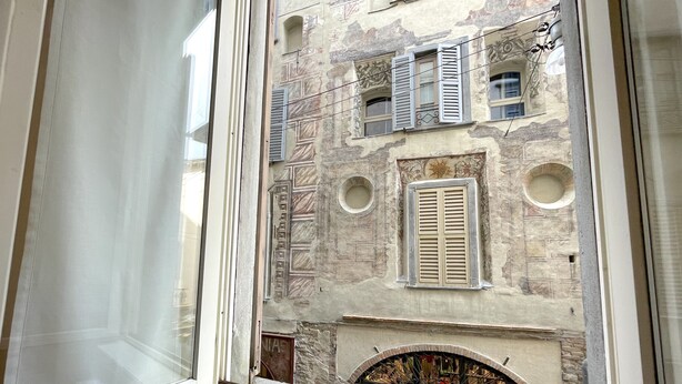 A view through a window of a building with multiple windows, some with shutters, and a circular window below. - Tower Gombito's Apartment (Bergamo)