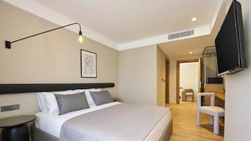 Une chambre d’hôtel moderne dotée d’un grand lit, d’une table de chevet, d’un téléviseur à écran plat fixé au mur et d’un couloir menant à un espace détente avec un fauteuil et une table.