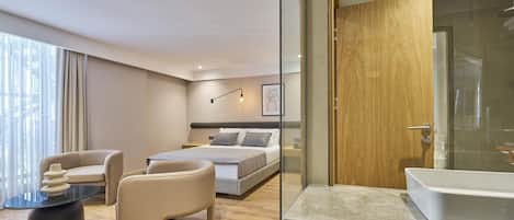 Una habitación de hotel moderna con una cama grande, una zona de estar con sofá y mesa de centro, cabina de ducha de vidrio y encimera de mármol en el baño.