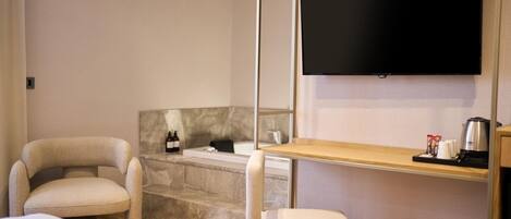 A modern hotel room with a bed, two chairs, a small table with a coffee maker, a flat-screen TV, and a marble-topped bathtub.