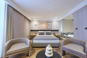 Una habitación de hotel moderna con una cama grande, dos sillones, una mesa de centro redonda y una decoración minimalista.