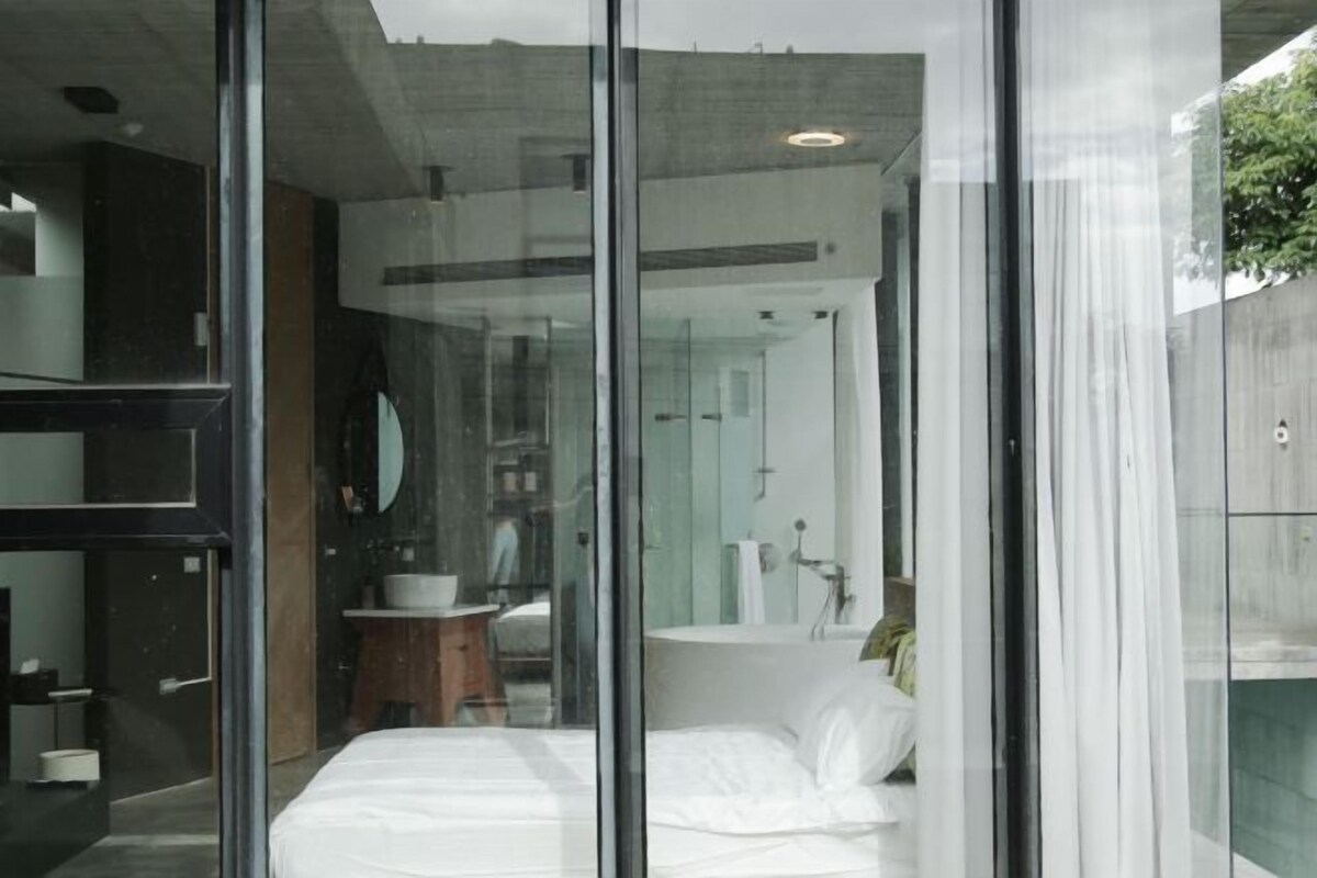 A modern bedroom with a glass wall, a bed, a bedside table, and a mirror.