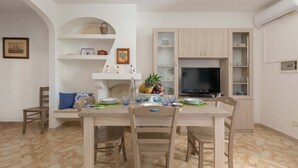 A dining area with a wooden table, chairs, and a fruit bowl. There is a TV on a stand and a shelf with decorative items.