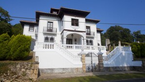 A white two-story building with a balcony on the second floor, a staircase leading to the entrance, and a gated front door. - Villa Victoria (AS)