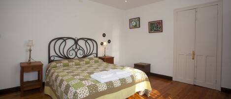 A bedroom with a wooden floor, a bed with a quilt, two bedside tables with lamps, a wardrobe, and two framed pictures on the wall.
