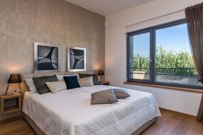 Ein Schlafzimmer mit einem großen Bett, zwei Nachttischen mit Lampen, einem Fenster mit Blick auf Grünflächen und abstrakter Kunst an der Wand.