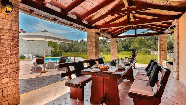 An outdoor patio with a wooden table and benches, surrounded by stone walls and a view of a swimming pool and greenery. - Villa Edera in Sveti Kirin (Sveti Kirin)