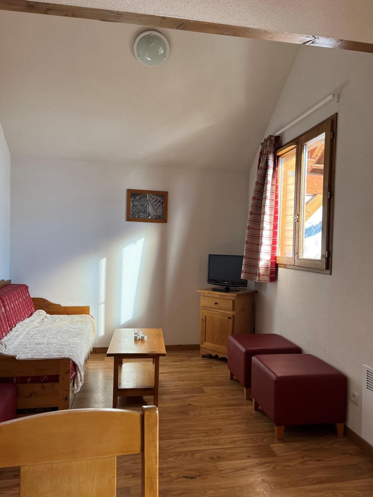 Ein gemütliches Zimmer mit einem Bett, einem Holztisch, einem Fernseher auf einer Holzkommode und einem Fenster mit Vorhängen.
