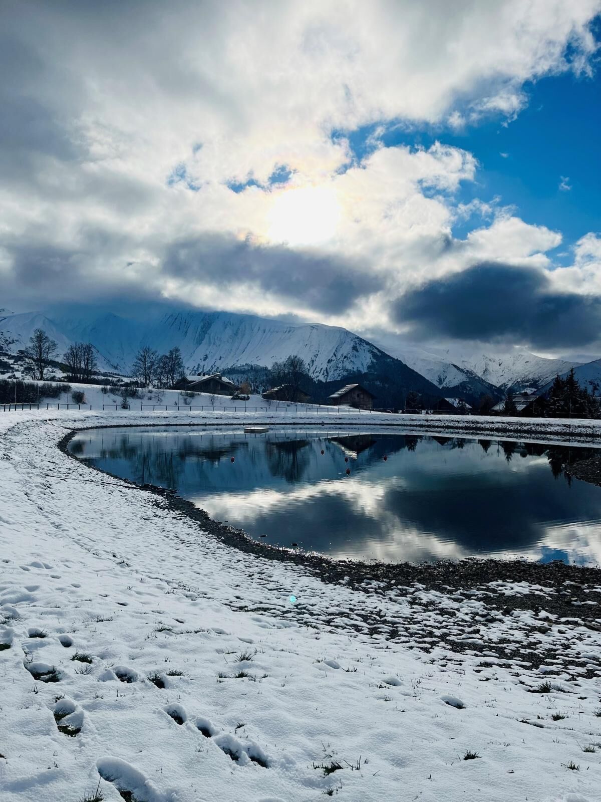 A snow-covered landscape with a calm lake reflecting the sky and mountains in the background.