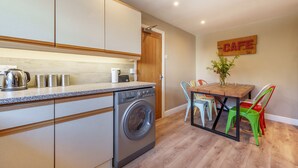 A compact kitchen with a washing machine, a coffee maker, and a dining area with a table and chairs. - Orrest Head Cottage,  located on the footpath to the famous Orrest Head walk. Perfect for dog walks. (Windermere)