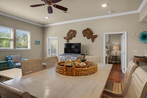 Un espace repas comprenant une grande table en bois, des chaises, un ventilateur de plafond et un téléviseur posé sur un meuble.