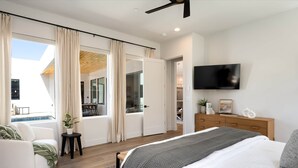 A modern bedroom with a large bed, a TV mounted on the wall, a wooden dresser, and a view of the outside through a sliding door.