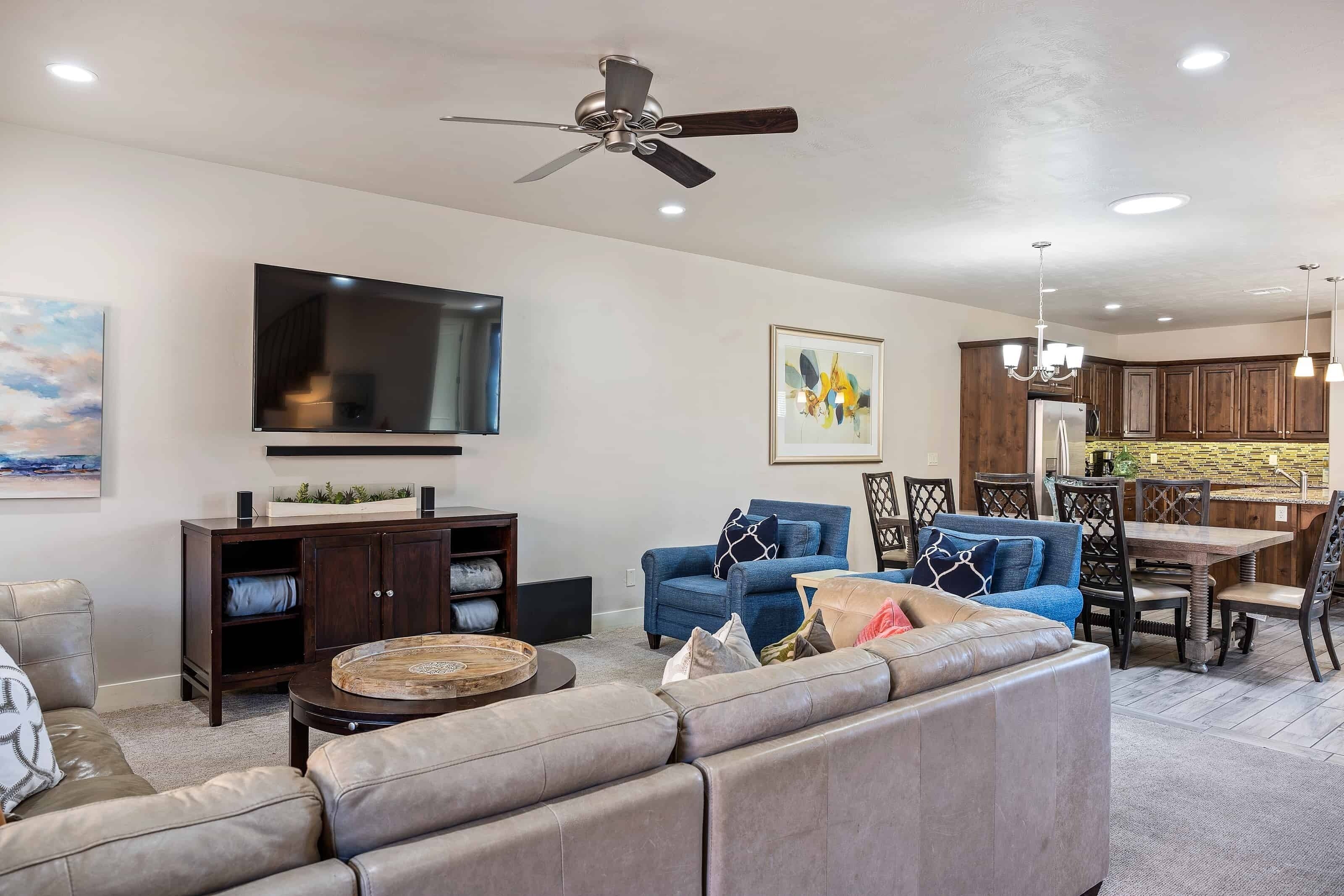 A modern living room with a sectional sofa, a coffee table, a TV mounted on the wall, and a dining area with a wooden table and chairs.
