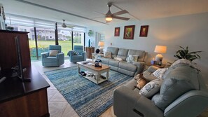 A spacious living room with a large window, a ceiling fan, and a grey sofa. - Beach-Pickleball-Golf-Tennis-Pools-Fitness Center-Restaurant all in one location (Fort Pierce)