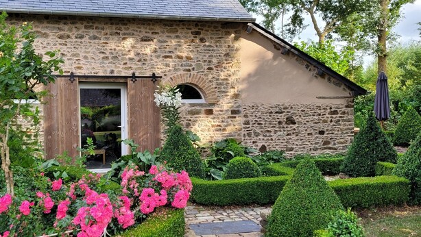 A stone building with a wooden door and window, surrounded by well-maintained hedges and flowering plants. - Maison de Campagne (Moulins)